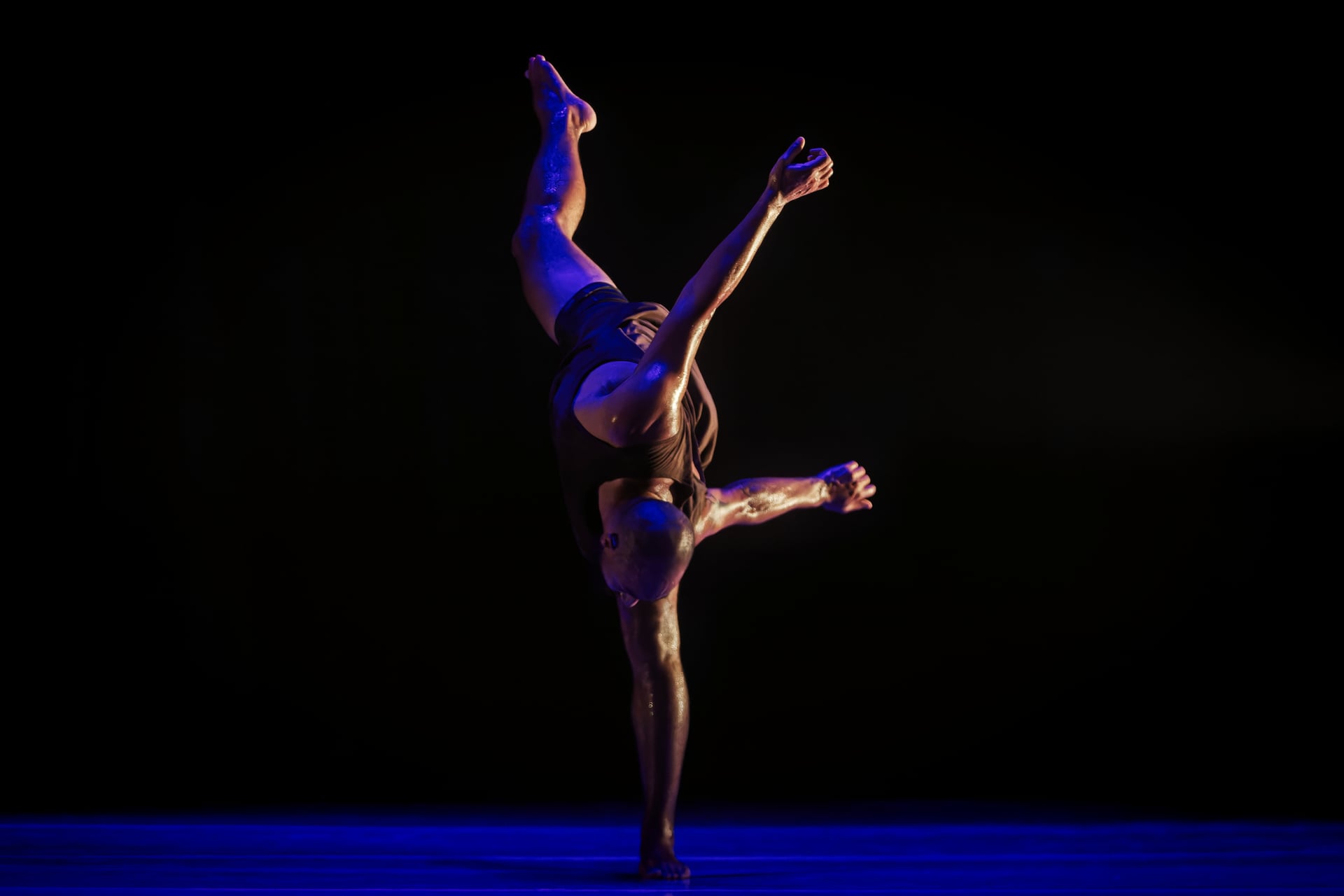 Intérprete en equilibrio sobre una pierna, con el torso inclinado y la otra pierna elevada bajo luz azul.